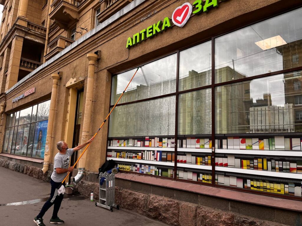 Мойщик очищает витрину аптеки телескопической штангой